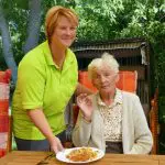 Betreuerin serviert Seniorin das Mittagessen im Freien