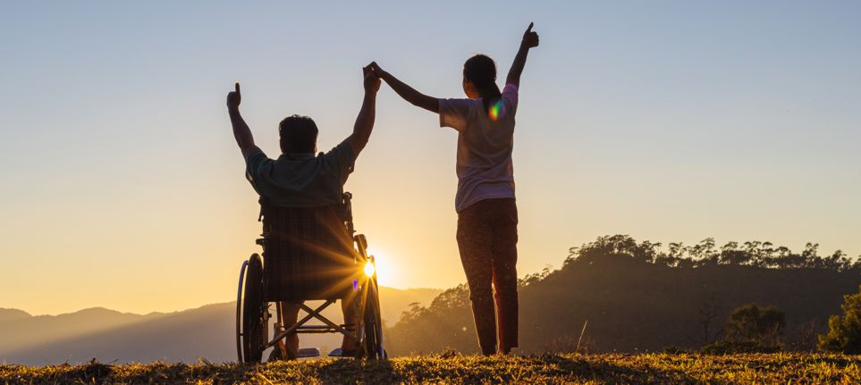 Rollstuhlfahrer und Frau im Sonnenuntergang erheben Hände in den Himmel