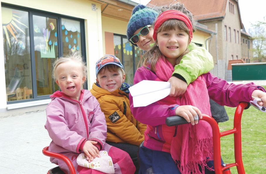 Mädchen mit Down Syndrom sitzt mit anderen Kindern auf einem Dreirad in der Kita